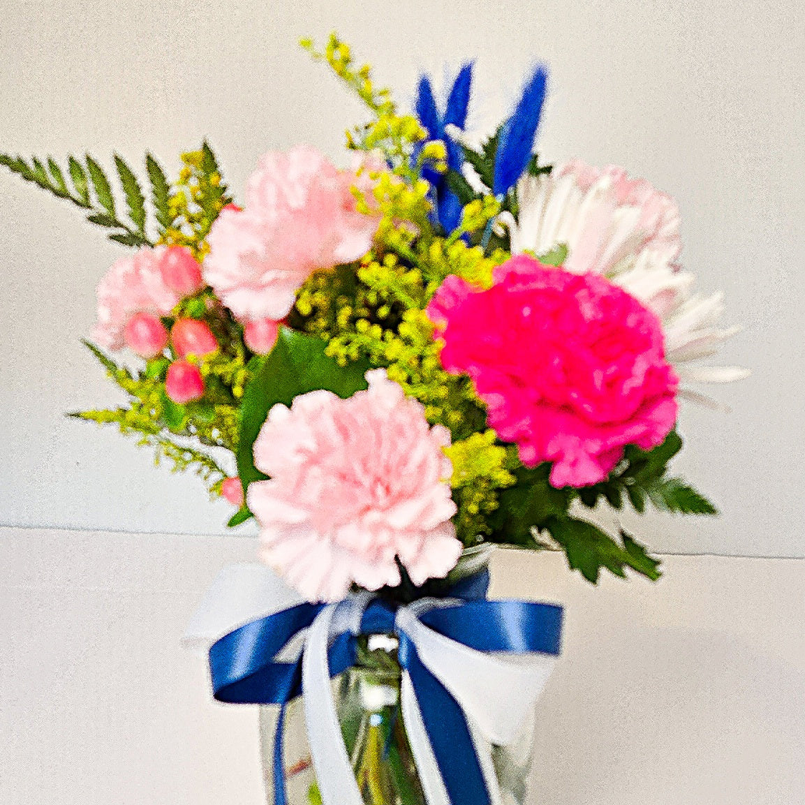 A vibrant flower bouquet in a clear glass jar vase, featuring pink carnations, a bright pink carnation, white daisies, green foliage, and blue ornamental grass, tied with a blue and white ribbon bow.