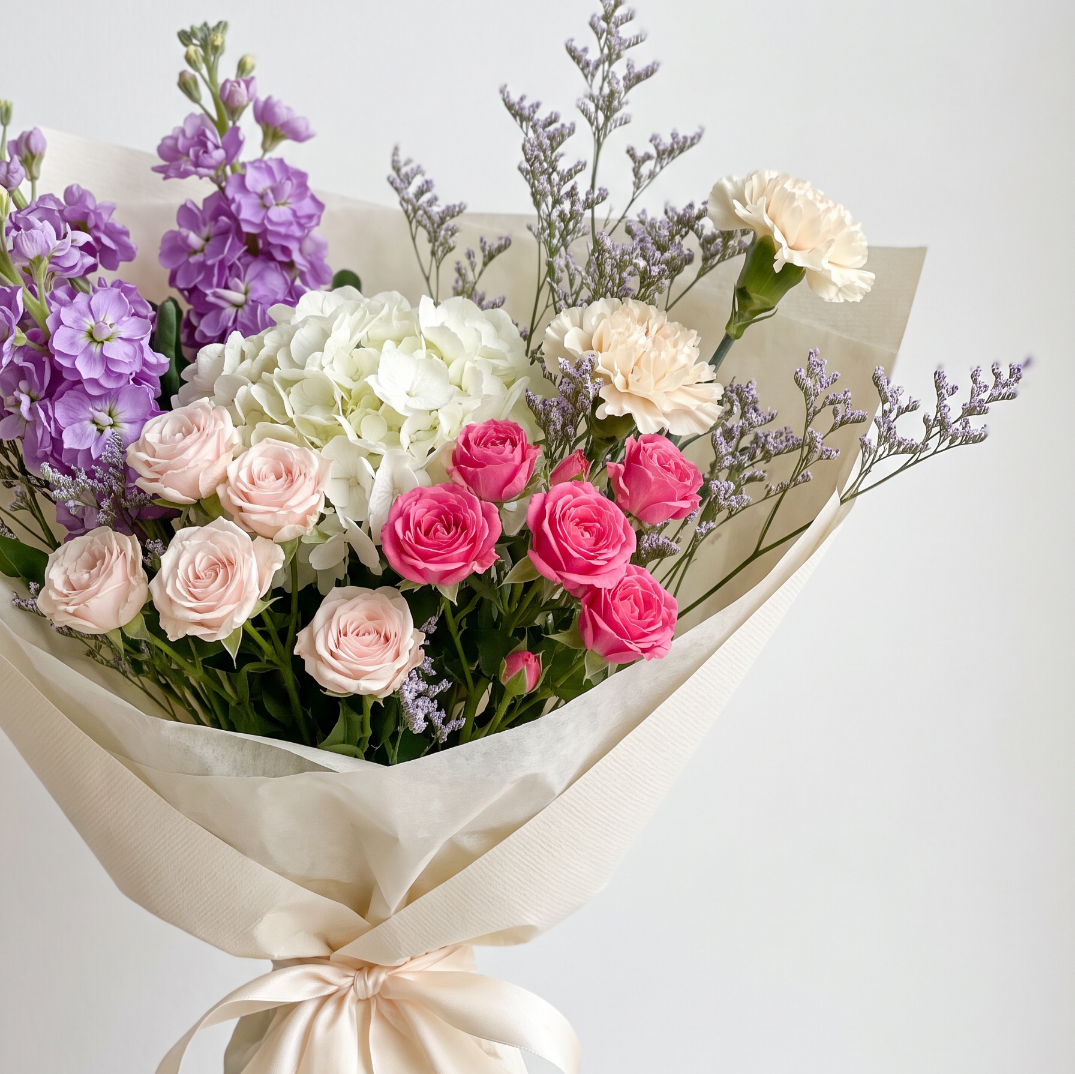 A beautifully arranged floral bouquet wrapped in cream-colored paper with a matching ribbon. The bouquet features a variety of flowers including purple stock, white hydrangea, pink and peach roses, a cream carnation, and light purple filler stems, set against a clean, light gray background. A luxury flower bpuquet from a Chilliwack florist.  Delivery in Sardis BC.
