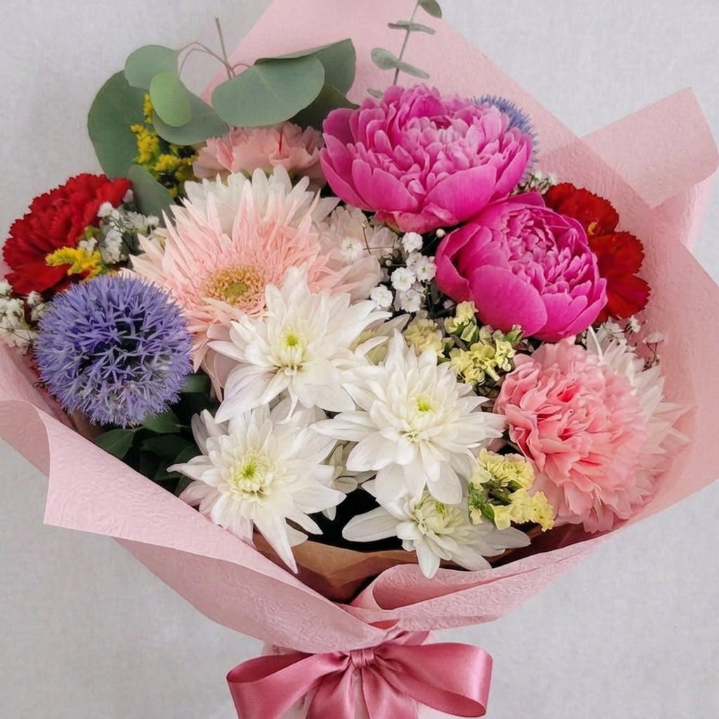 A vibrant mixed flower bouquet wrapped in peach and beige paper, featuring bright pink and red carnations, white chrysanthemums, pink gerbera daisies, a purple globe flower, and green eucalyptus leaves on a light gray background.