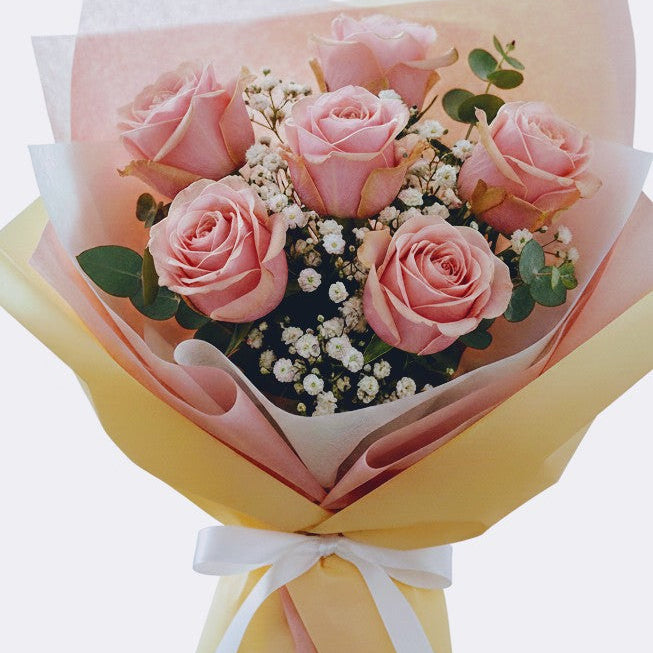 A bouquet of six pink roses wrapped in pastel pink and yellow paper with a white ribbon, set against a plain light gray background.